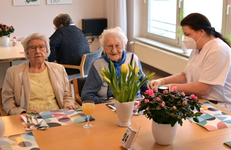 Bewohnerinnen des Convenio Gelsenkirchen am Tisch mit Frühlingsblumen, betreut von einer Pflegekraft